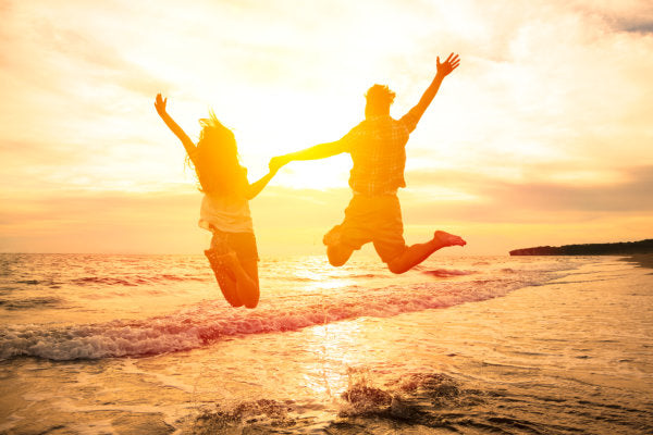 Paar springt freudig in die Luft vor Sonnenuntergang am Meer – Symbol für Leichtigkeit und Lebensfreude