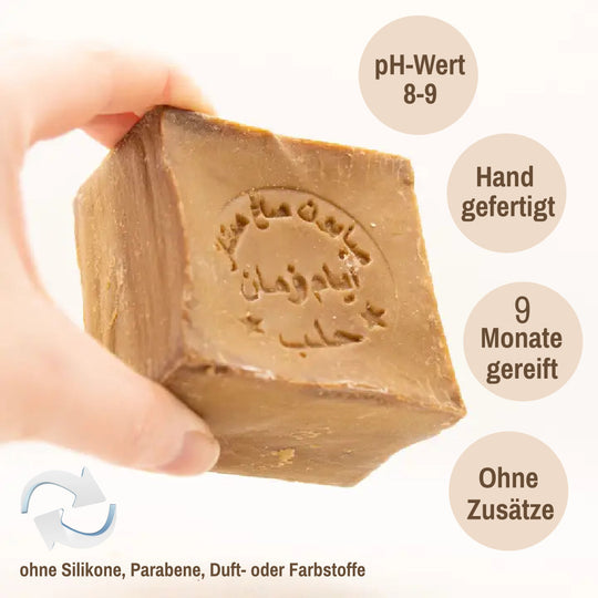 Hand, die eine Aleppo Seife mit geprägter arabischer Schrift hält; daneben sichtbare Texte „pH-Wert 8–9“, „Hand gefertigt“, „9 Monate gereift“, „Ohne Zusätze“ sowie unten „ohne Silikone, Parabene, Duft- oder Farbstoffe“.
